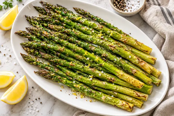 Roasted asparagus spears with browned tips on a white platter beside a lemon wedge